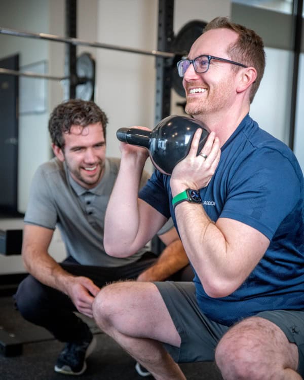 Man performing kettlebell exercise with physiotherapist supervising in gym.