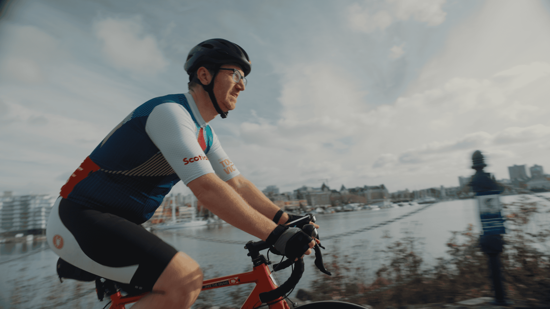 Male cyclist riding along waterfront with city skyline in background.