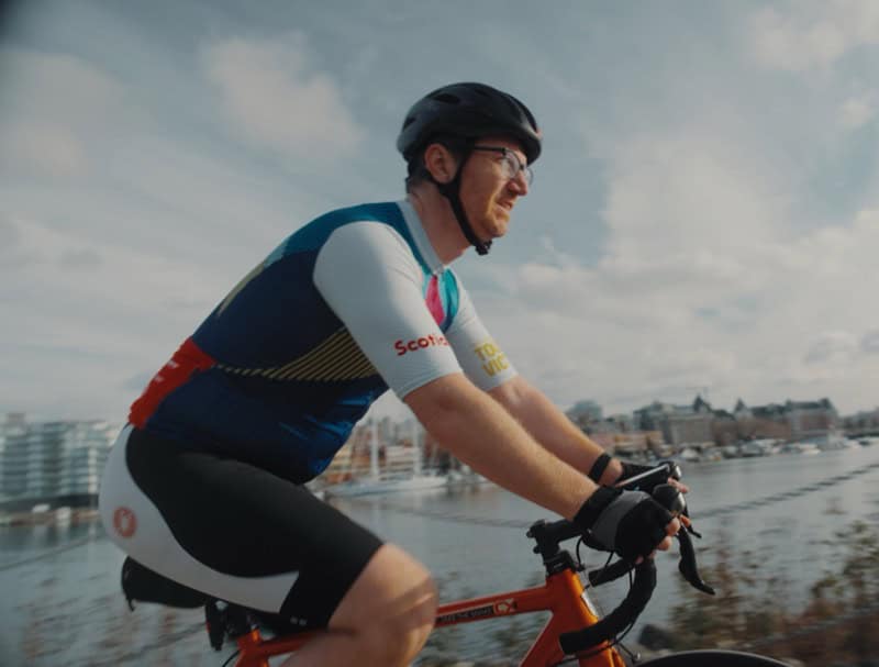Woman cycling along waterfront with city skyline in background.